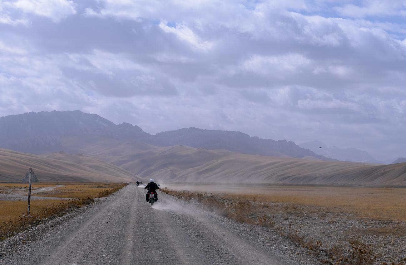 A motorcyclist rides a dusty gravel road through sweeping rolling hills and mountains on the Silk Route.