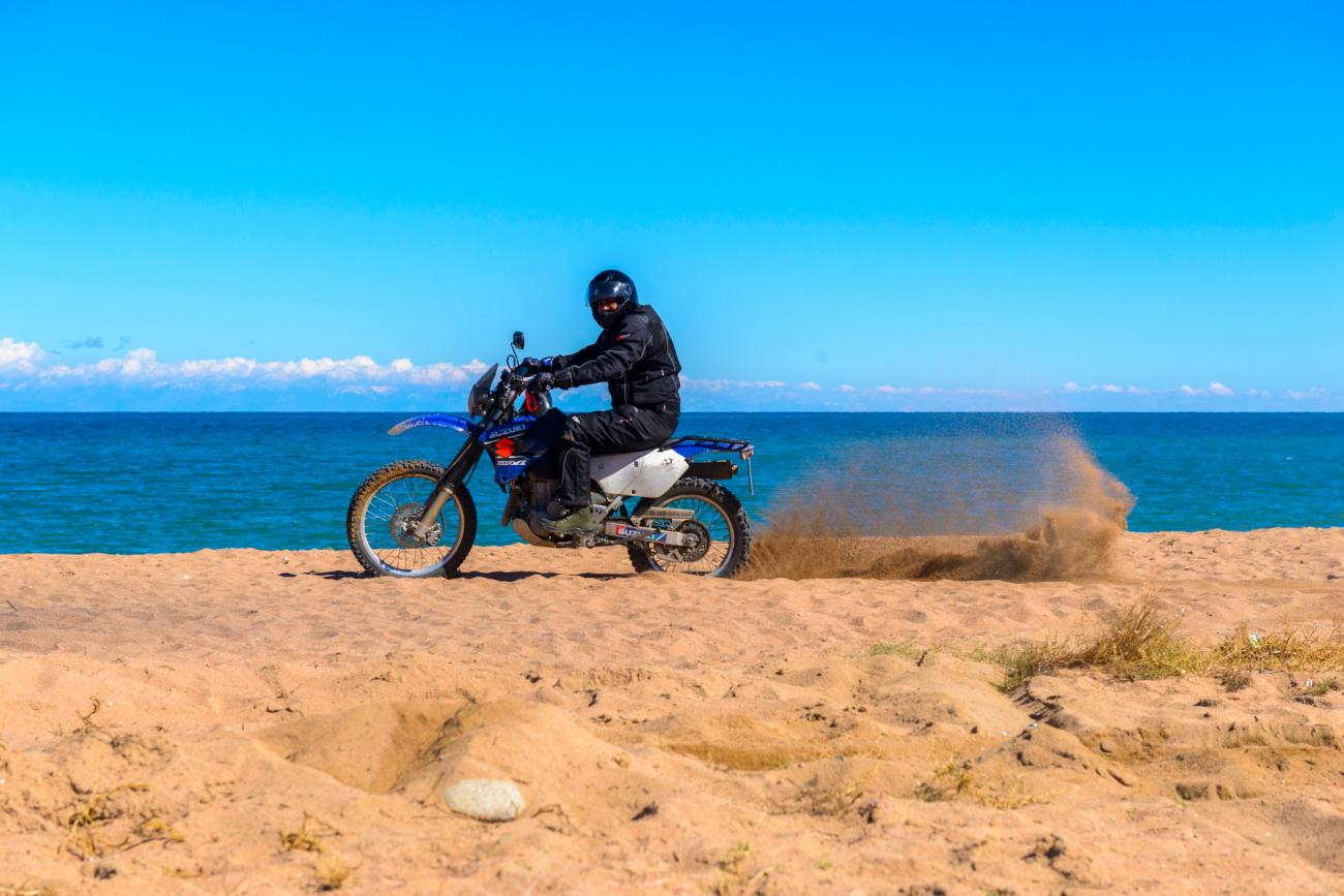A motorcyclist kicks up sand while riding along the shore of Issyk-Kul Lake with turquoise water stretching to the horizon.