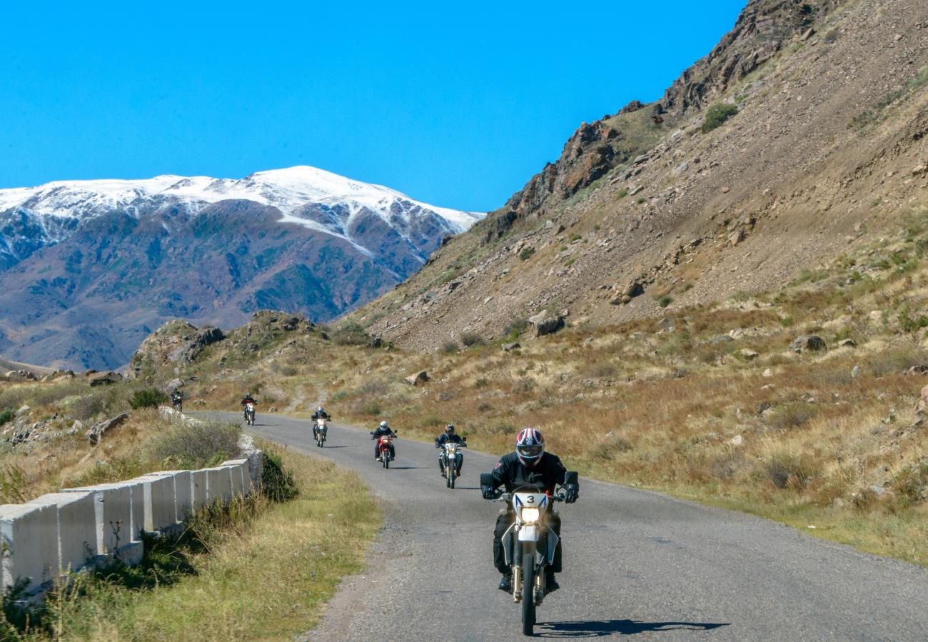 Motorcyclists ride up a winding mountain road with snow-capped peaks towering in the background along the Silk Route.