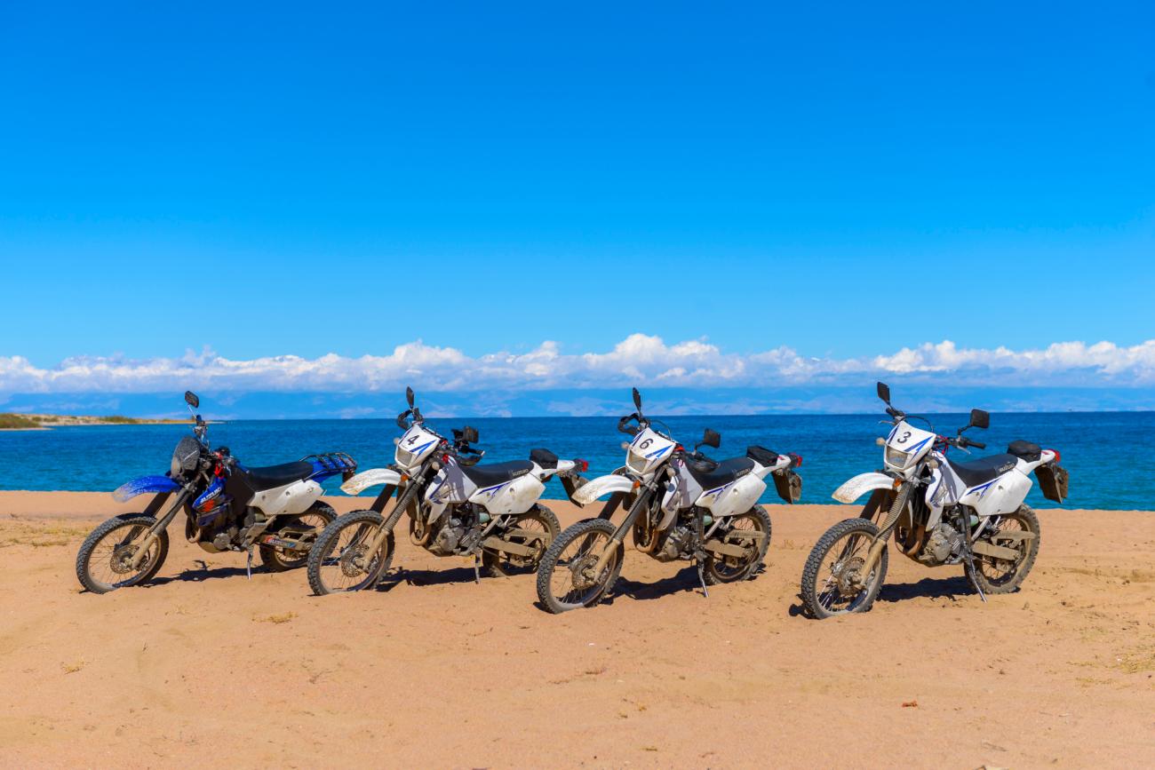 Five adventure motorcycles parked in a row on a sandy beach beside the blue waters of Issyk-Kul Lake in Kyrgyzstan.