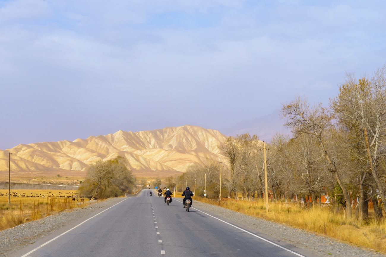 Motorcyclists ride a straight road toward dramatic eroded sandstone mountains in the Kyrgyz countryside.