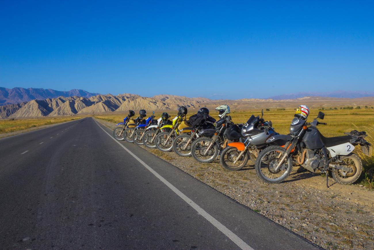 A row of adventure motorcycles parked along a highway with dramatic eroded ridgelines stretching into the distance.