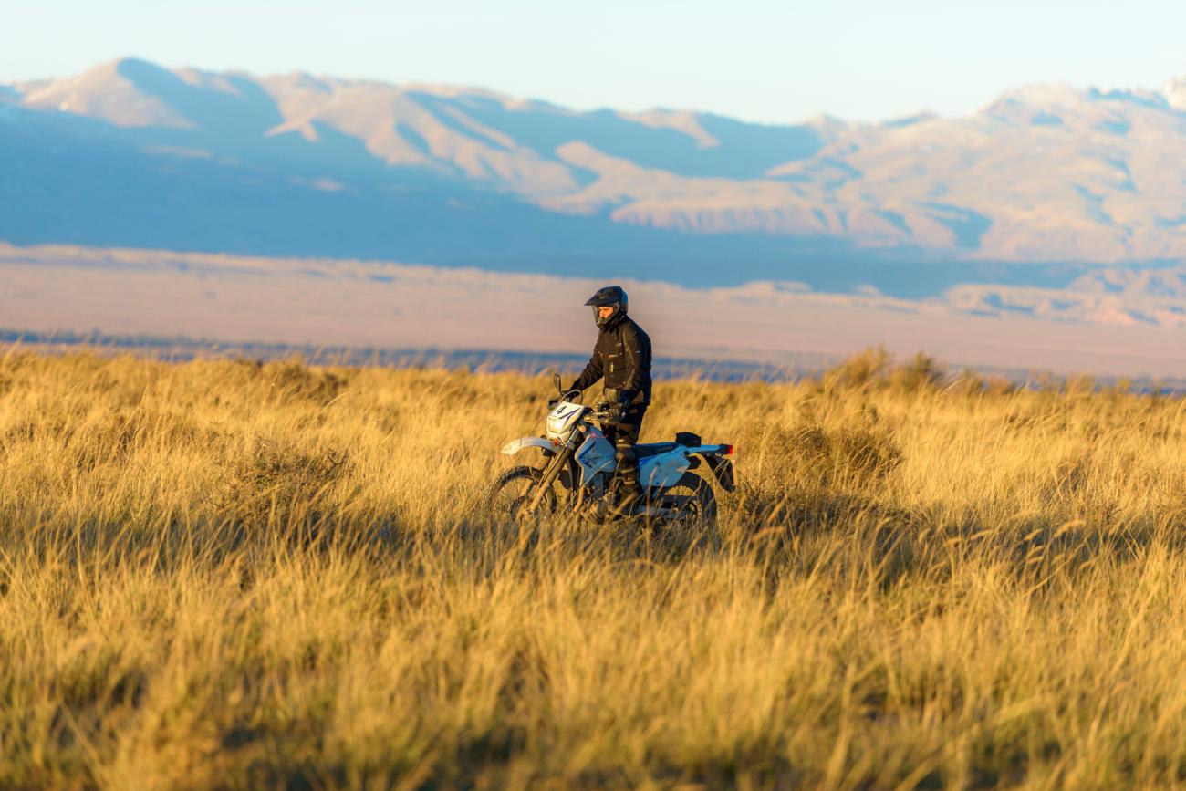 A lone motorcyclist stands beside his bike in golden steppe grass at sunset with snow-dusted mountains in the distance.