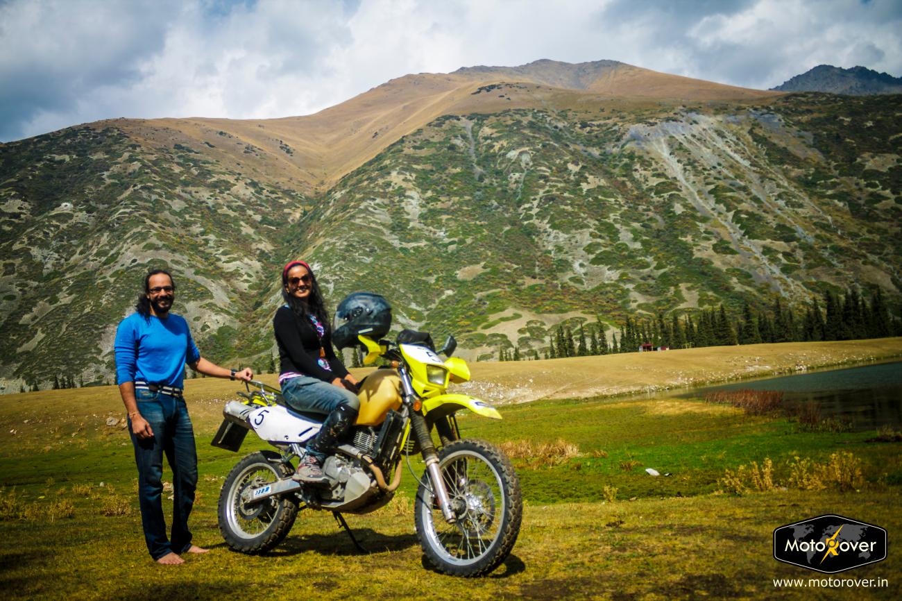 Two riders pose with a yellow motorcycle on a green highland pasture surrounded by tall Kyrgyz mountains.
