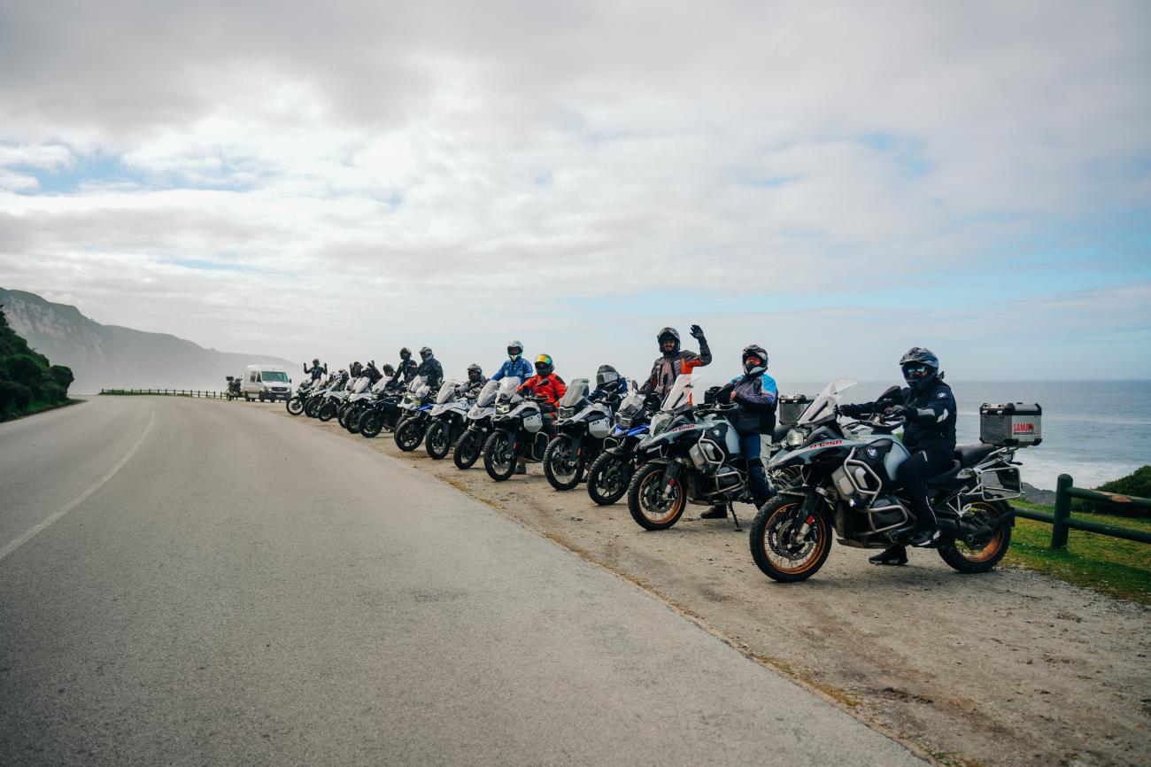 Large group of BMW adventure motorcycles lined up along a misty coastal road with cliffs and ocean in the background.
