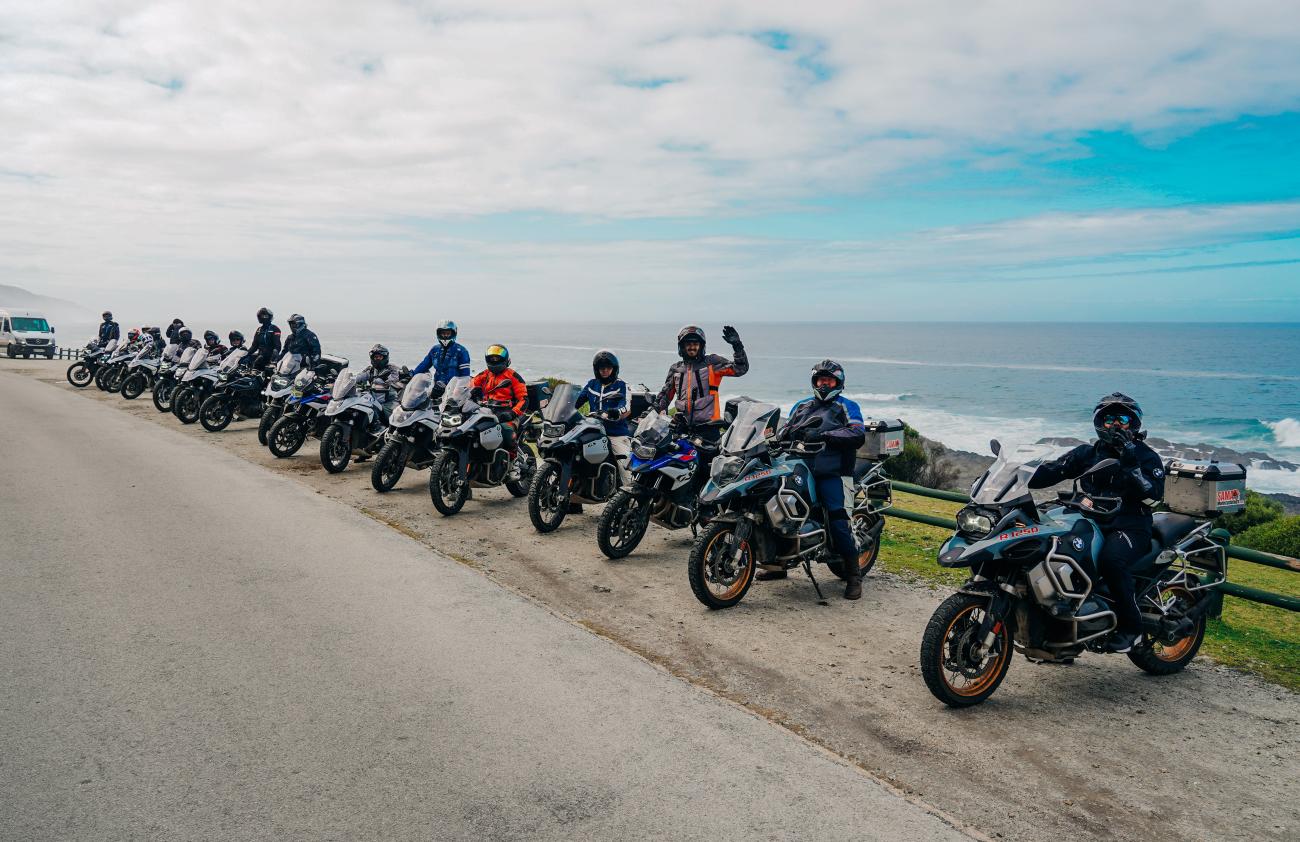Long line of BMW adventure motorcycles and riders parked at a coastal viewpoint along the Garden Route with ocean waves below.