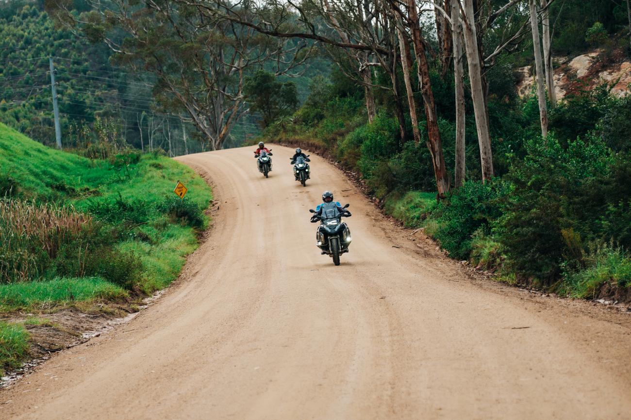 Three motorcyclists riding along a winding gravel road through green hills and eucalyptus trees in South Africa.