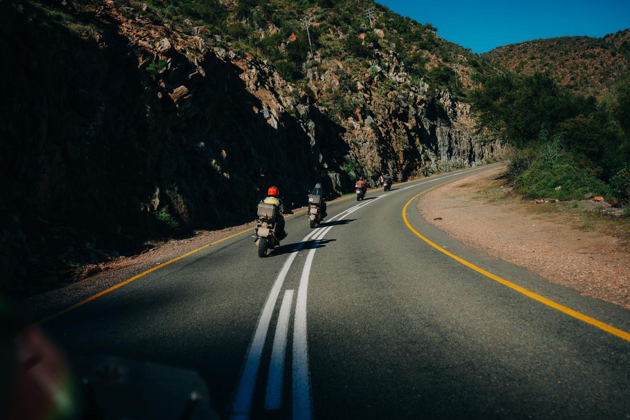 Motorcyclists riding through a dramatic rock-cut mountain pass with sweeping curves in the South African countryside.