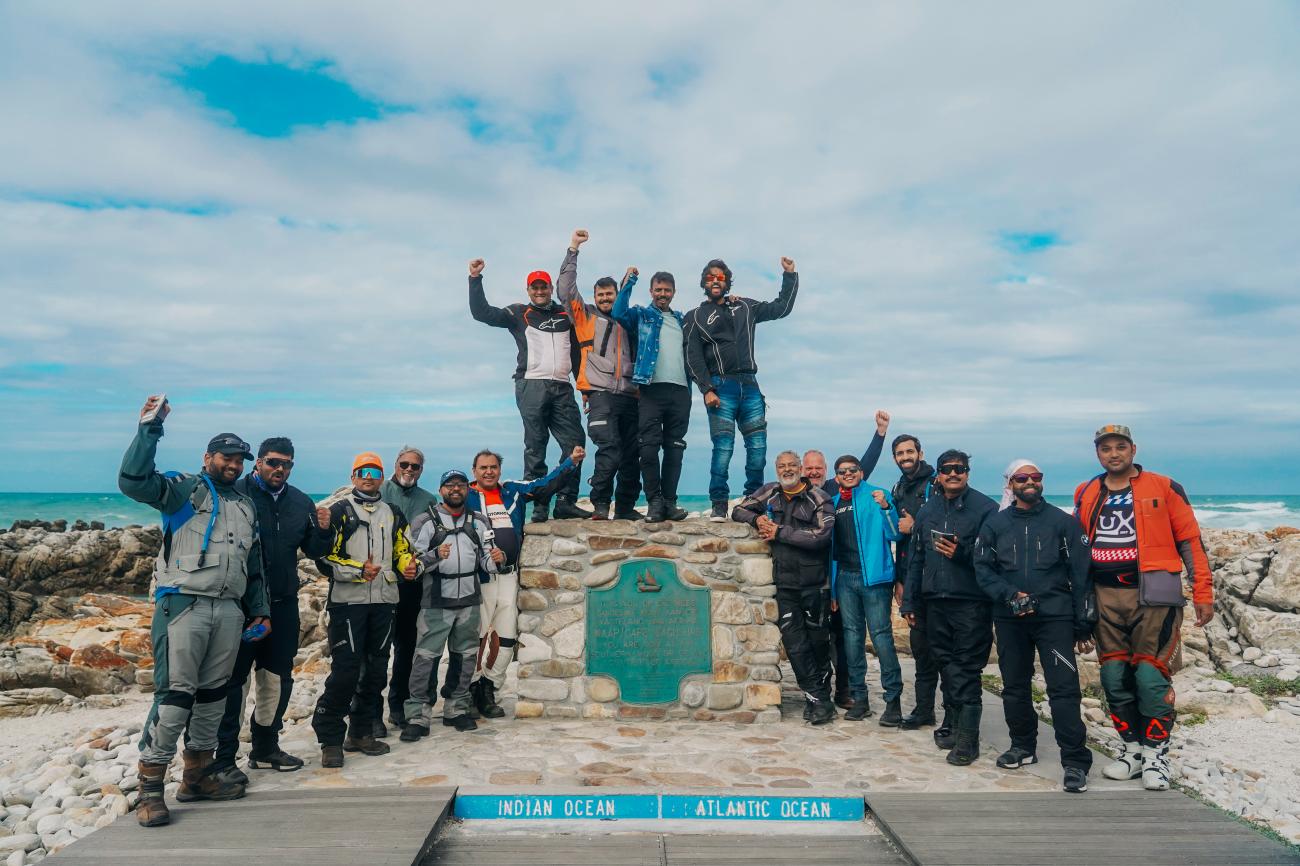 Motorcycle tour group celebrating at the Cape Agulhas marker where the Indian and Atlantic Oceans meet, the southernmost point of Africa.