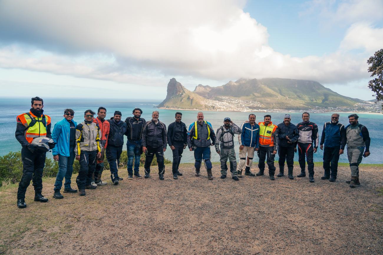 Motorcycle tour group posing for a photo overlooking Hout Bay with dramatic cloud-covered mountains behind them.