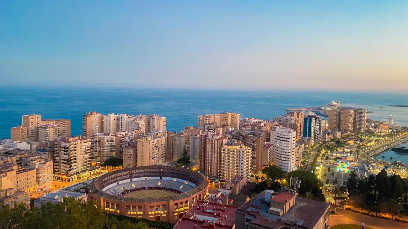 Aerial dusk view of Malaga showing the bullring, city skyline, and Mediterranean coastline.