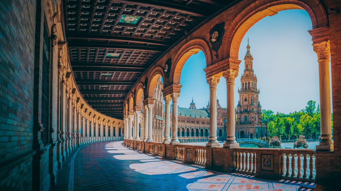 Sunlit colonnade with ornate arches and tower view inside the Plaza de Espana in Seville.