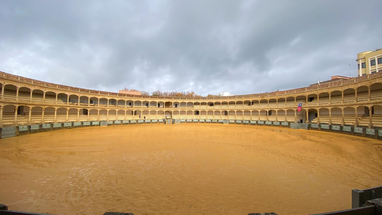 Interior of the historic Ronda bullring with its sandy arena and tiered arched galleries under an overcast sky.