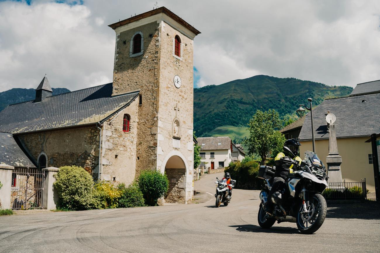 BMW GS adventure motorcycles parked beside a medieval stone church with a bell tower in a French Pyrenees village.