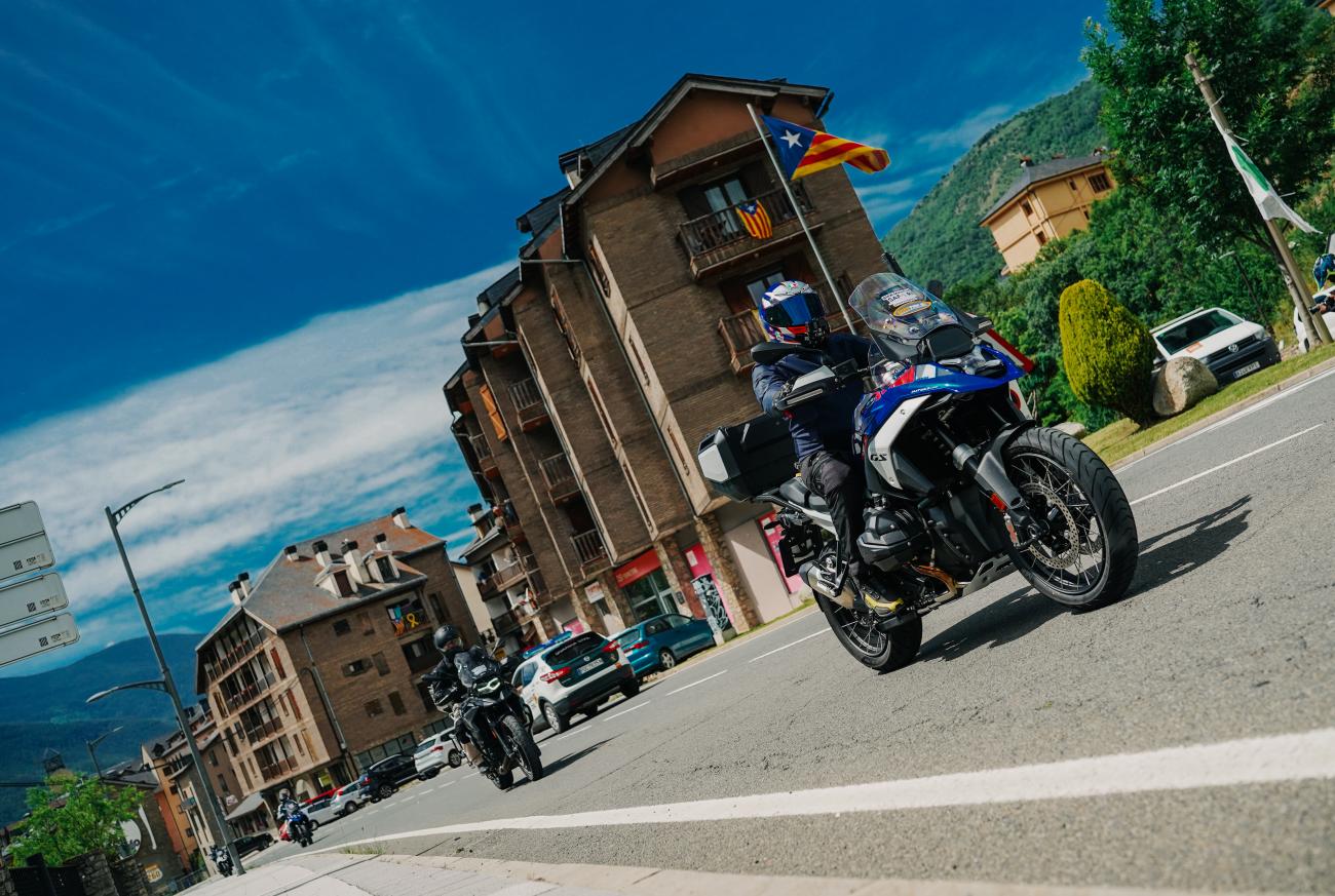 Adventure motorcycle riding through a Catalan Pyrenees village with traditional stone buildings and a Catalan flag.