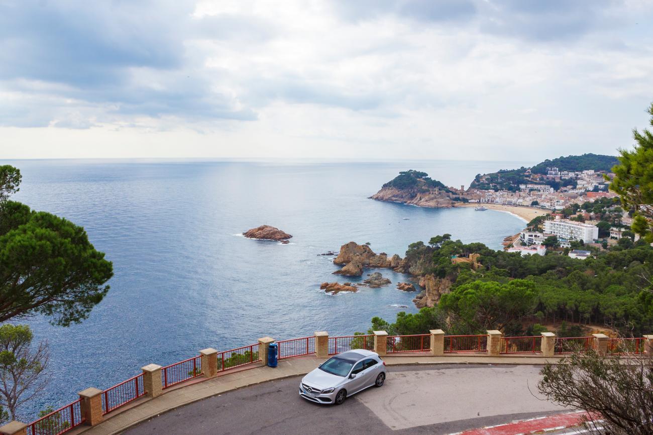 White Mercedes parked at a Costa Brava viewpoint overlooking rocky coves and the seaside town of Tossa de Mar.