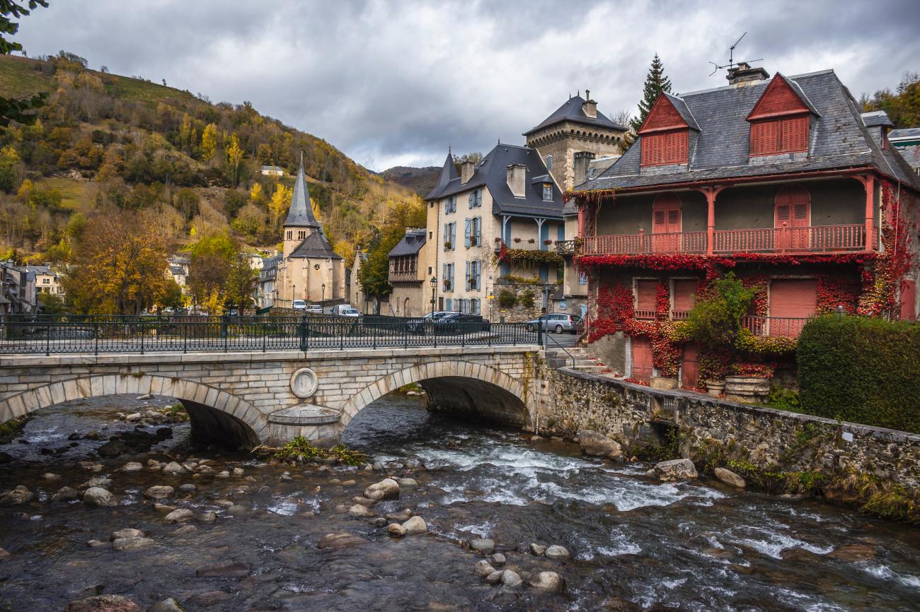 Stone bridge over a rushing river in a picturesque French Pyrenees town with ivy-covered red timber balconies and a church spire.