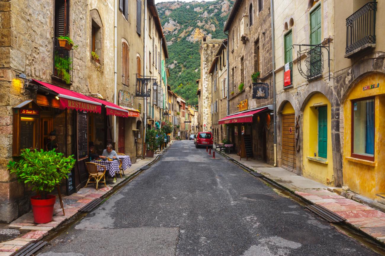Narrow cobblestone street lined with colorful restaurants and medieval stone buildings in a southern French village.
