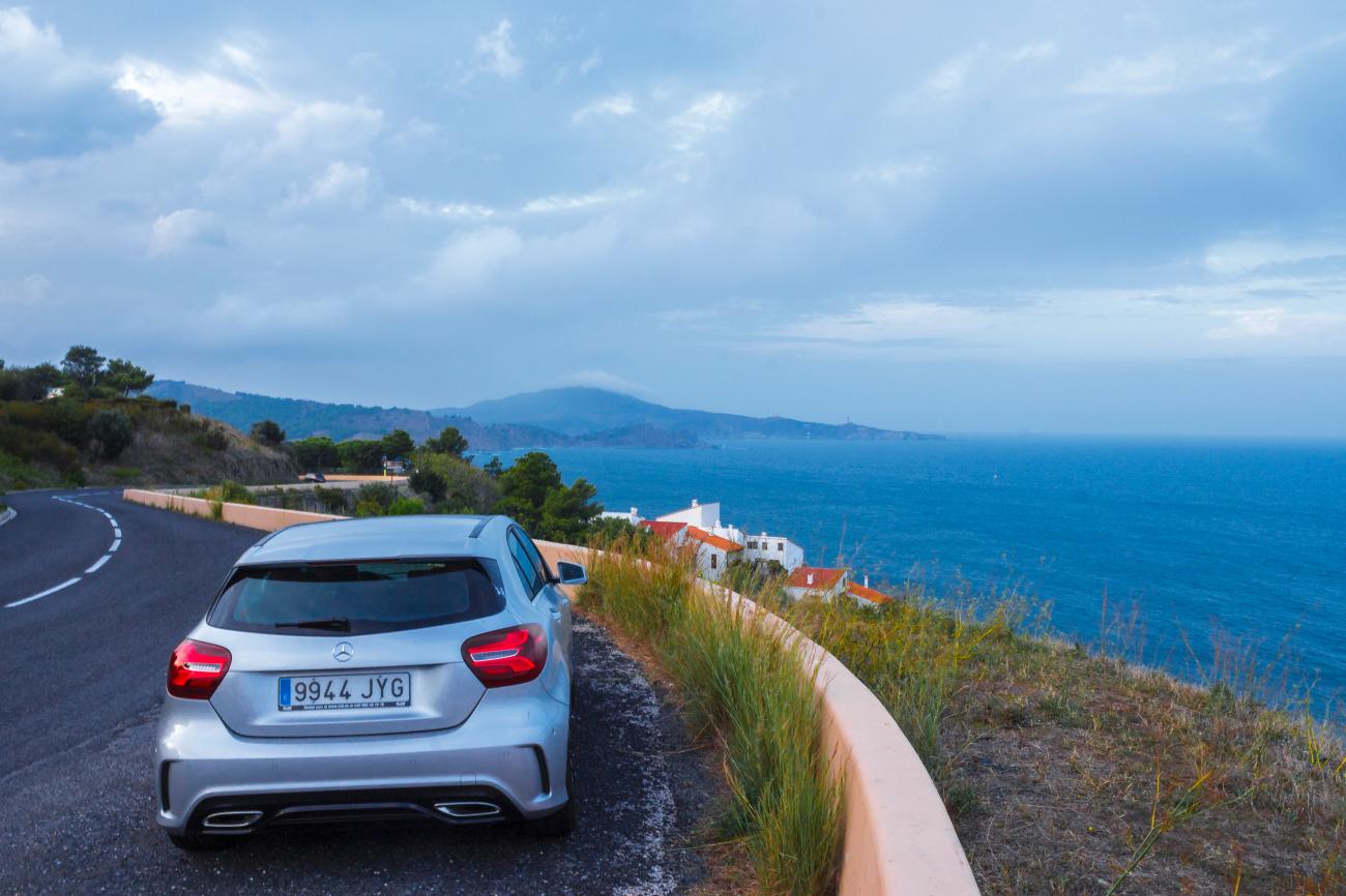 Silver Mercedes-Benz driving along a winding Mediterranean coastal road with blue sea and distant headlands.