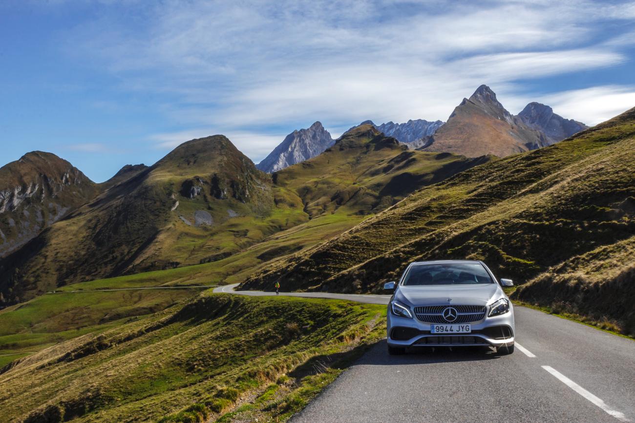 Silver Mercedes-Benz approaching on a smooth mountain road surrounded by lush green Pyrenees peaks under a blue sky.