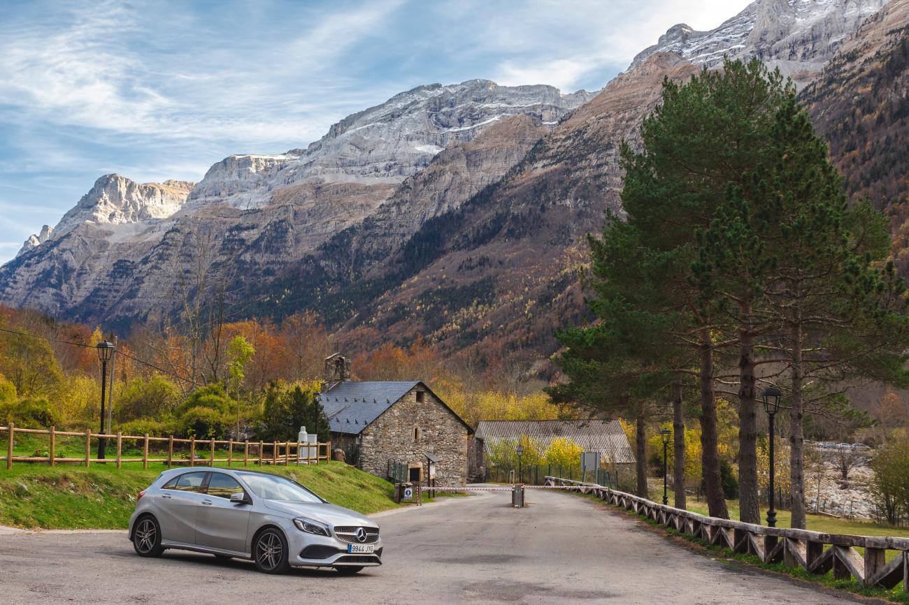 Silver Mercedes parked beside a stone village with towering limestone cliffs of the Pyrenees rising behind autumn foliage.