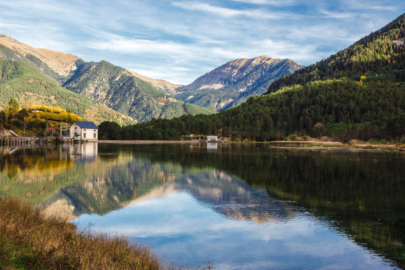 Calm Pyrenees lake reflecting forested mountains and a small stone building on its shore in autumn.