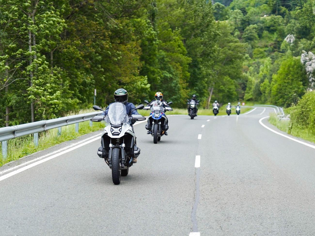 A group of adventure motorcyclists riding together on a tree-lined European road.