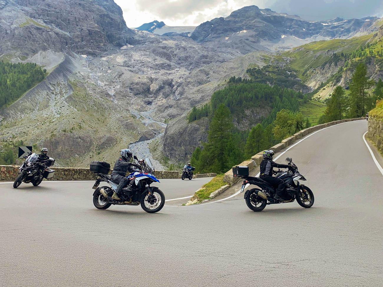Motorcyclists navigating a hairpin turn on Stelvio Pass with a dramatic glacier and rocky alpine landscape behind them.