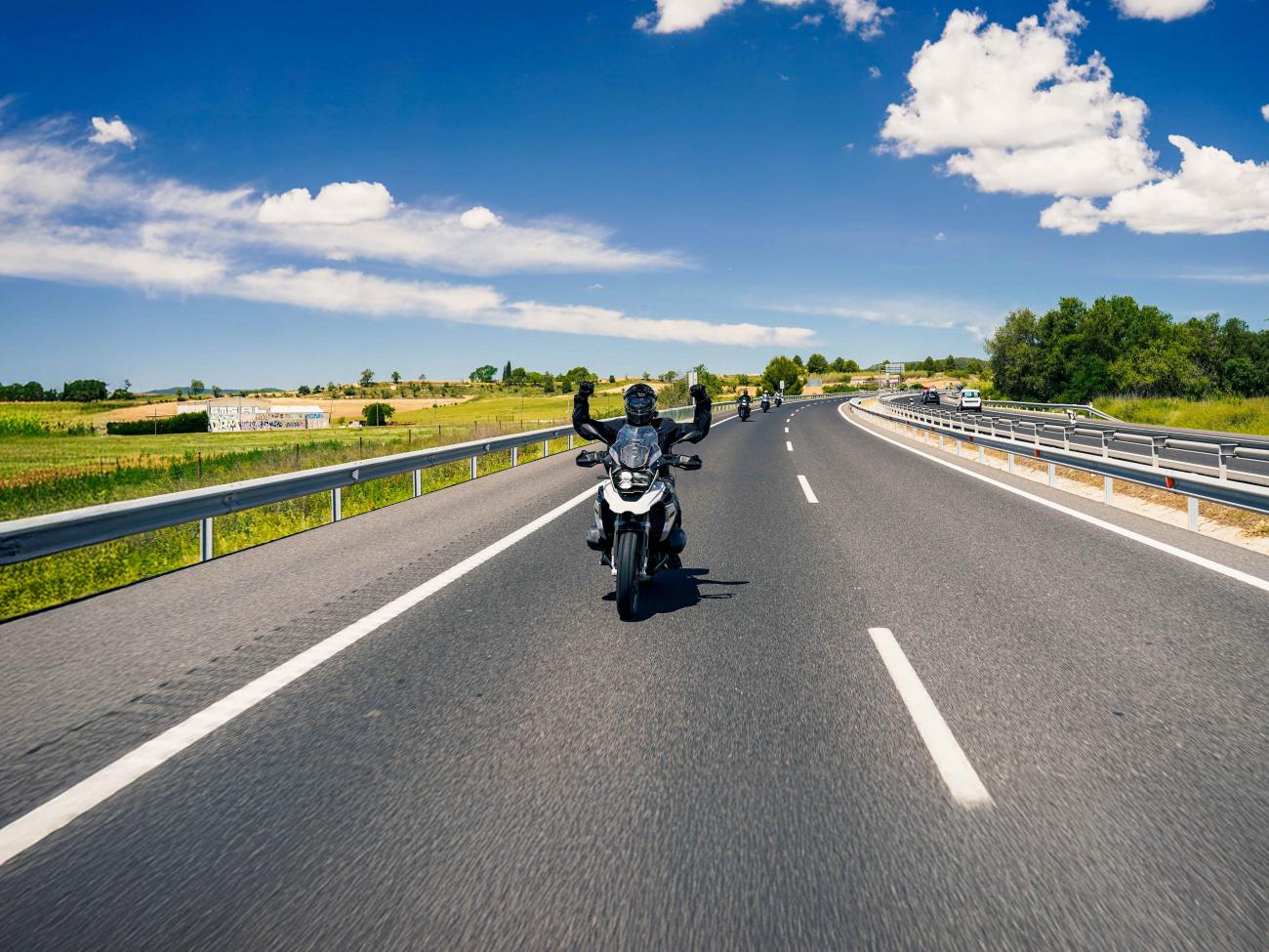 A motorcyclist raises both fists in celebration while riding on an open European highway under blue skies.