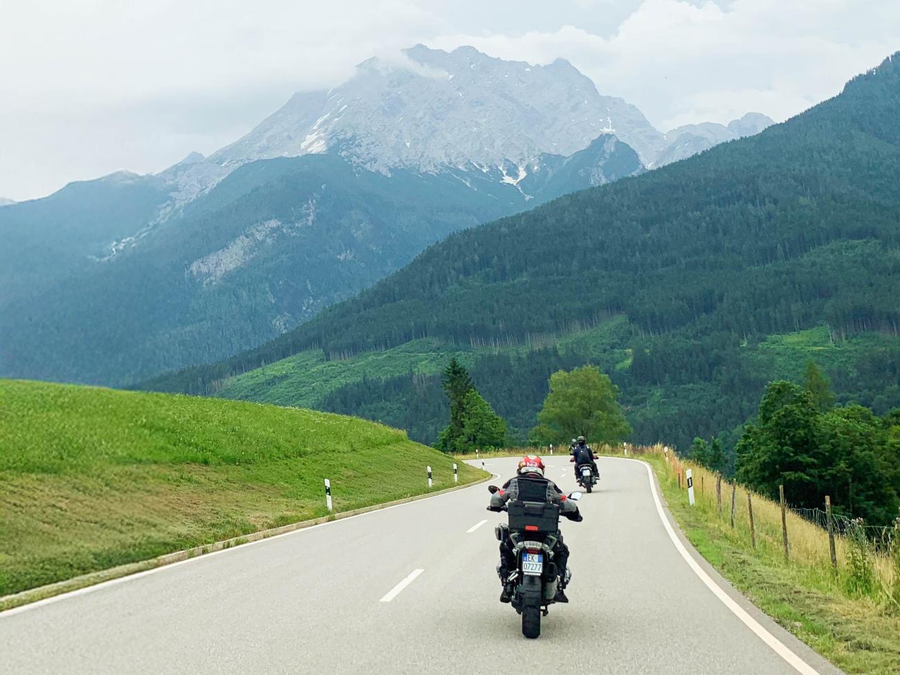 Motorcyclists riding in formation on an alpine road with snow-capped peaks and green meadows ahead.